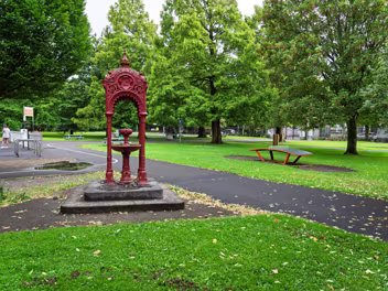 THE TAIT FOUNTAIN A tall, ornate cast-iron drinking fountain, redin colour, with a filigree dome and intricate detailing, standing in a...